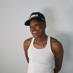 Person wearing a white tank top and San Junipero cap with a red logo on a plain background