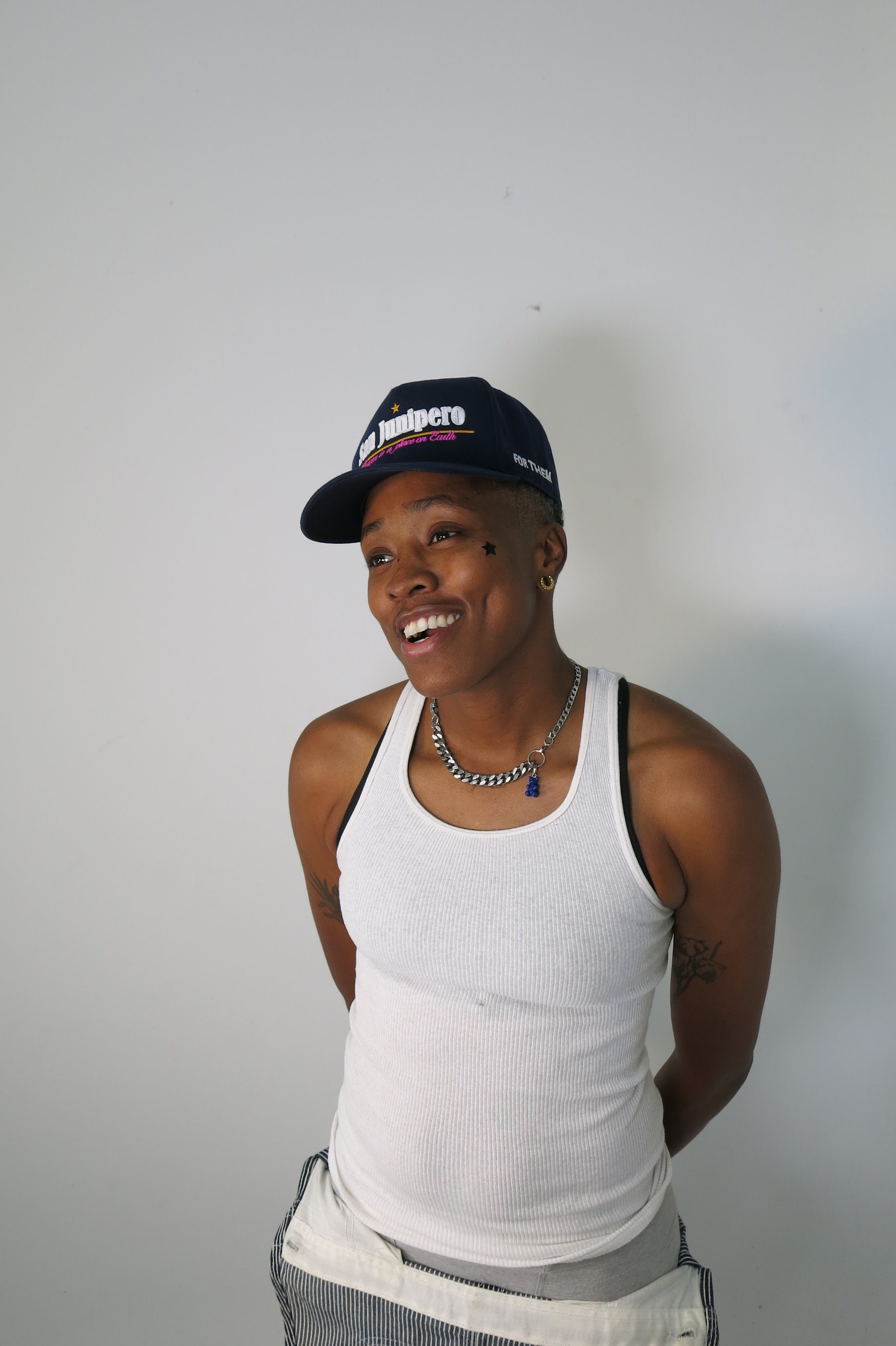 Person wearing a white tank top and San Junipero cap with a red logo on a plain background